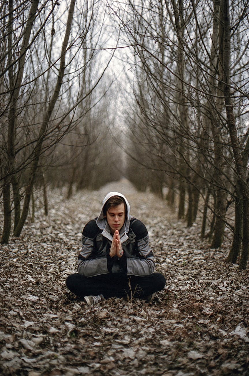 man wearing gray black zip hoodie jacket praying in between black tree during daytime