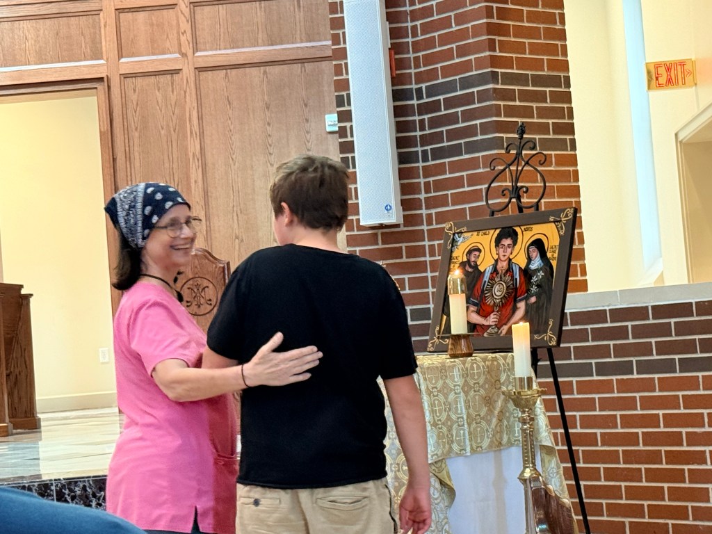 A woman in a pink shirt and headscarf warmly greets a boy in a black shirt near an altar with religious icons and candles, in a church setting.