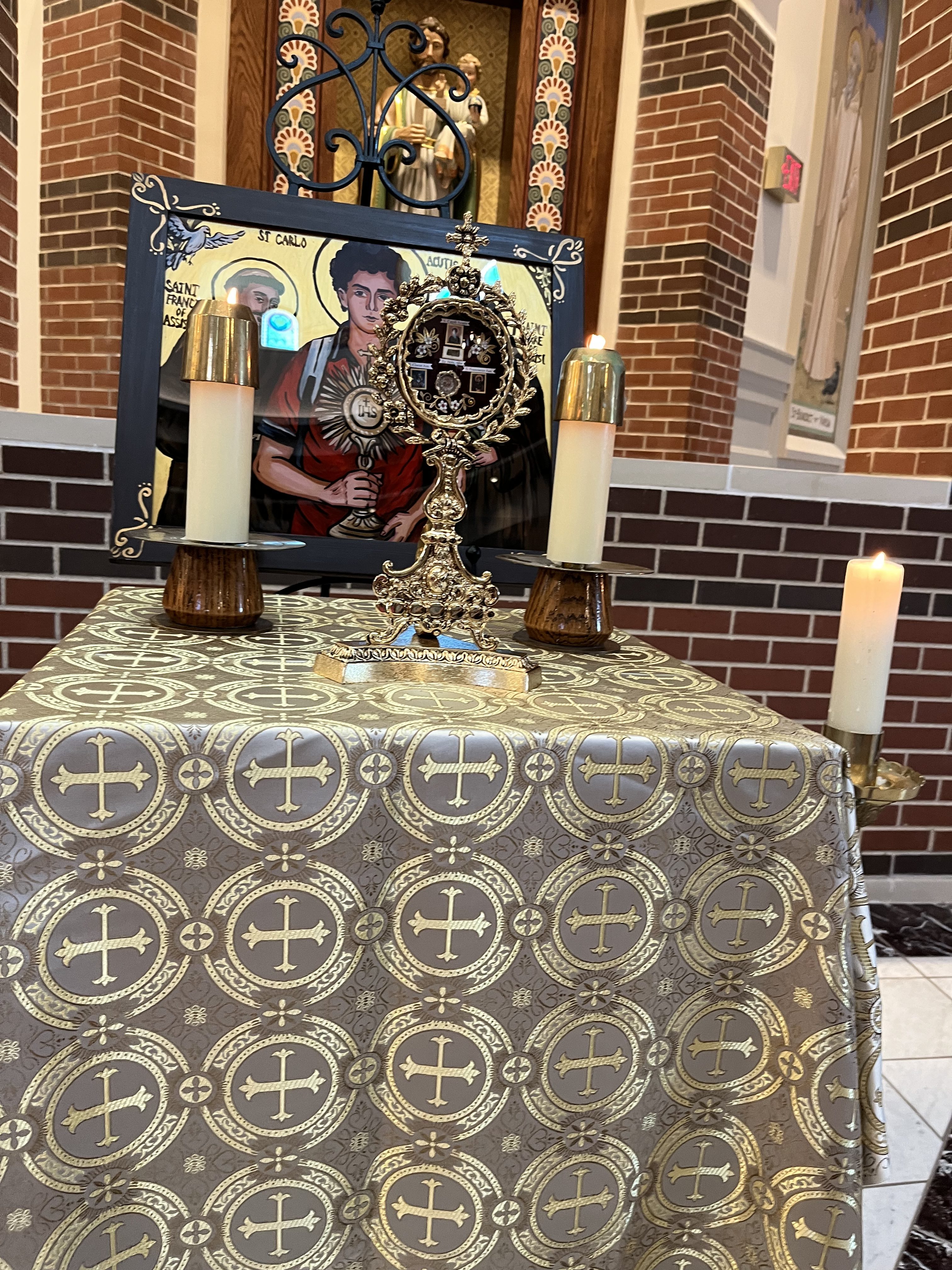 A table in a church adorned with a decorative cloth featuring golden crosses, a gold ornate monstrance with candles on either side, and a framed image of Saint Carlo.